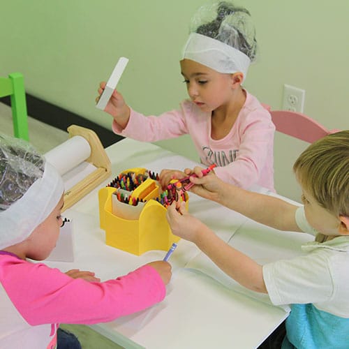 Group of kids sitting at a little table coloring with crayons, they have shower caps on for the lice treatment oil